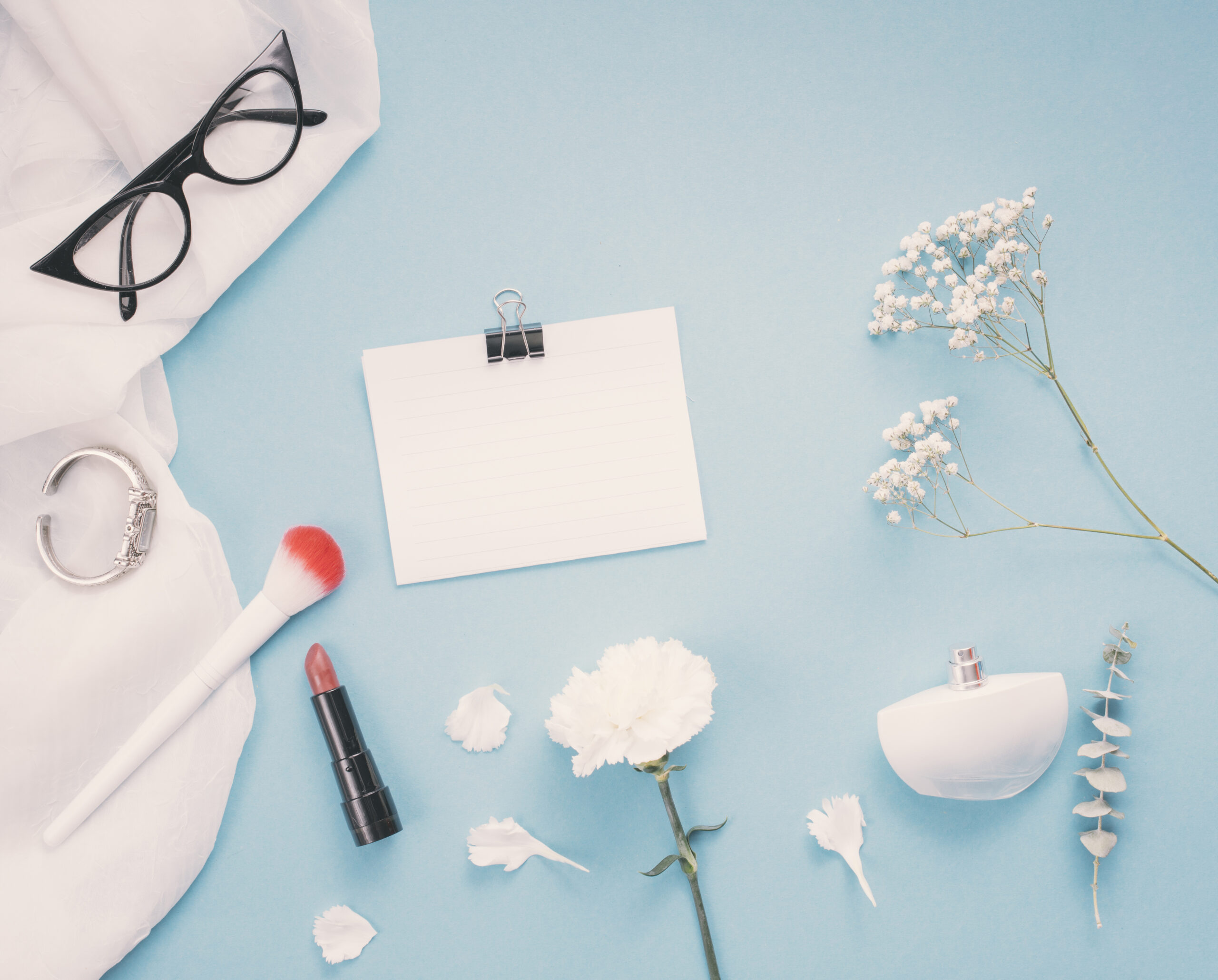 Flat lay com produtos de maquiagem, perfume e flores sobre fundo azul, representando testes e regularização de cosméticos no Brasil.