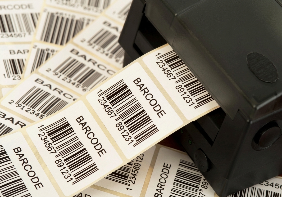 Close de uma impressora térmica profissional imprimindo etiquetas brancas com código de barras e o texto "BARCODE"