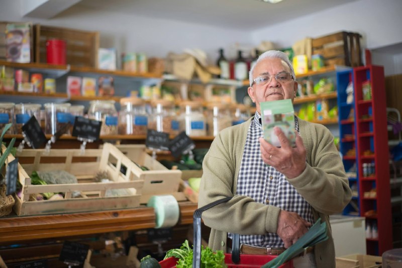 Homem sênior, de óculos, lendo atentamente o rótulo de uma caixa em um mercado orgânico, segurando uma cesta de compras. Ao fundo, prateleiras de madeira desfocadas com potes de vidro e vegetais frescos. Ilustração para post sobre rotulagem de alimentos ANVISA.