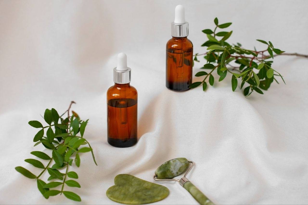 Dois frascos de fitocosméticos de vidro âmbar com conta-gotas, cheios de óleo vegetal, posicionados sobre linho branco com ramos verdes e um kit de massagem de jade.