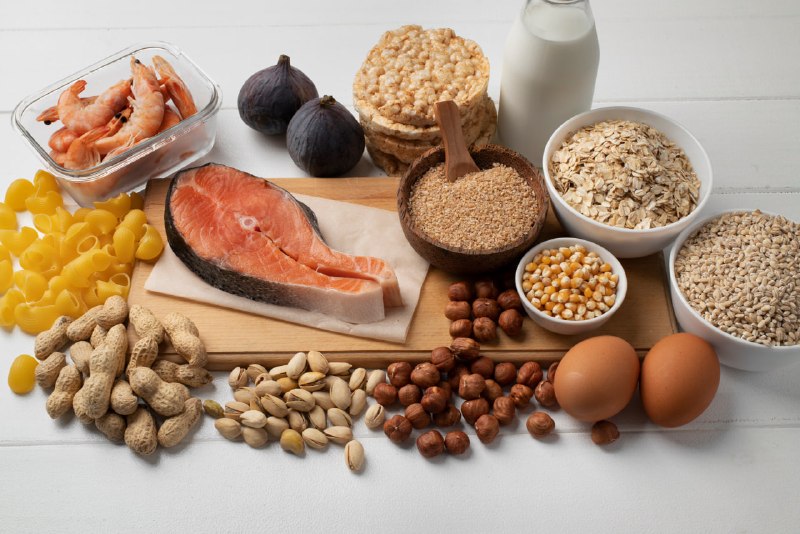 Diversos alimentos que são alérgenos comuns dispostos em uma mesa branca, incluindo amendoim, camarão, ovos, leite, peixe (salmão) e grãos contendo glúten.