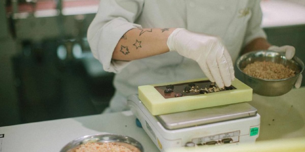 Confeiteiro com luvas e dólmã pesando barra de chocolate artesanal em balança de precisão durante processo de controle de qualidade.