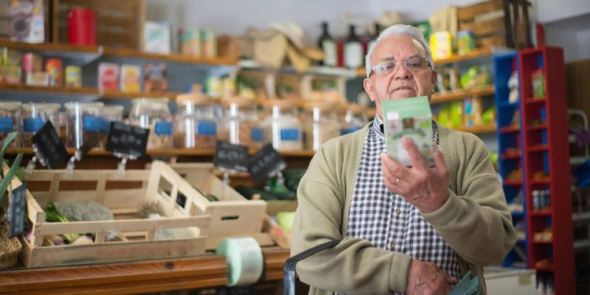Homem sênior, de óculos, lendo atentamente o rótulo de uma caixa em um mercado orgânico, segurando uma cesta de compras. Ao fundo, prateleiras de madeira desfocadas com potes de vidro e vegetais frescos. Ilustração para post sobre rotulagem de alimentos ANVISA.