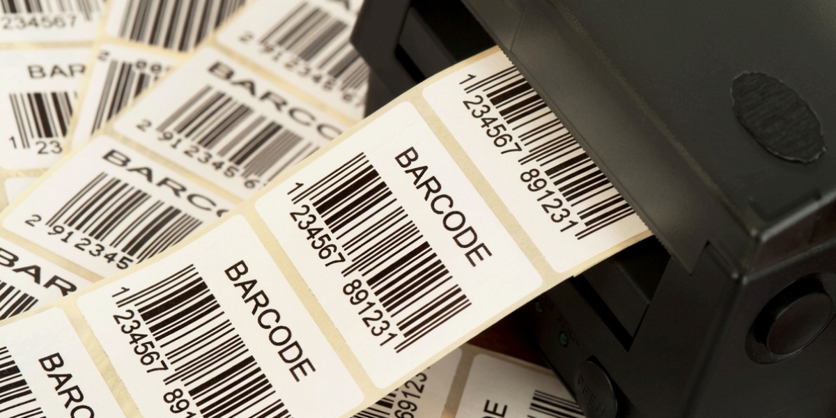Close de uma impressora térmica profissional imprimindo etiquetas brancas com código de barras e o texto "BARCODE"