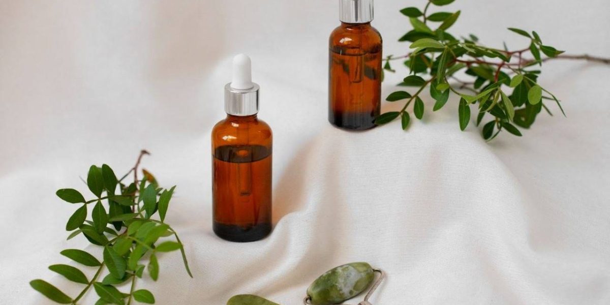 Dois frascos de fitocosméticos de vidro âmbar com conta-gotas, cheios de óleo vegetal, posicionados sobre linho branco com ramos verdes e um kit de massagem de jade.