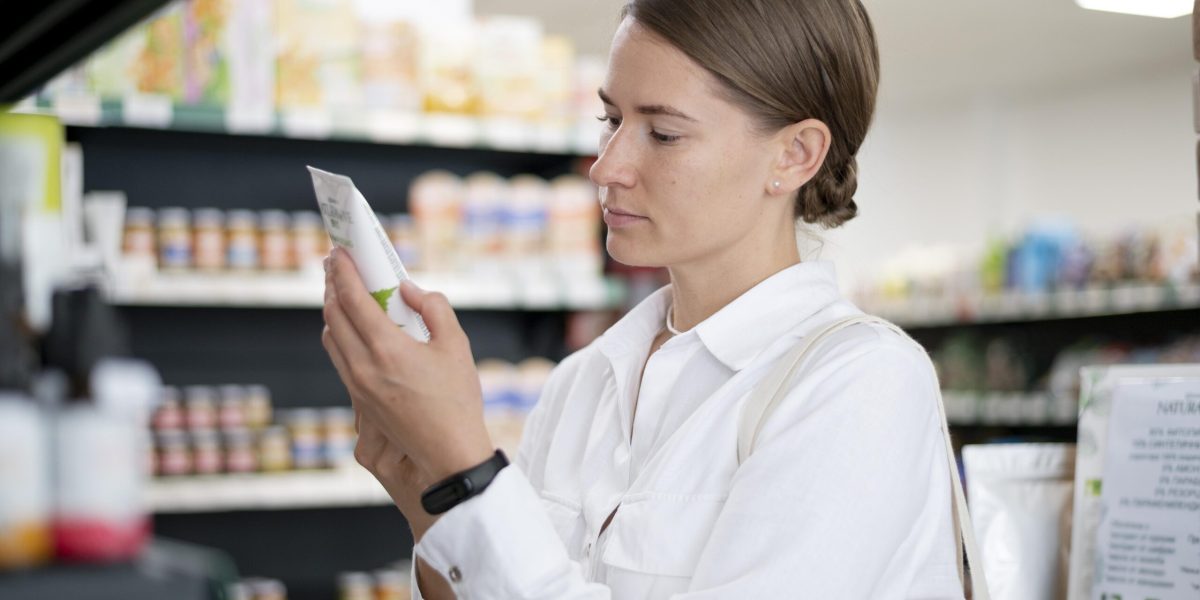 Mulher em supermercado analisando rótulo de produto, ilustrando a verificação de autenticidade de cosméticos, suplementos e medicamentos.