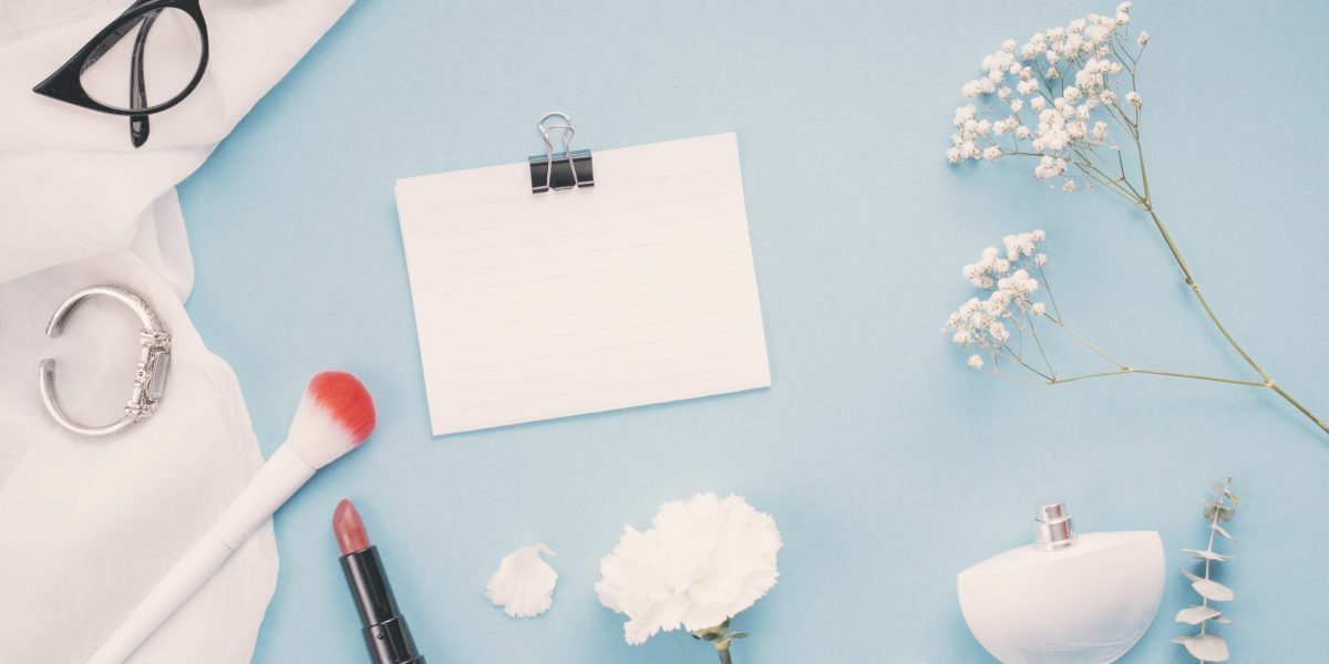 Flat lay com produtos de maquiagem, perfume e flores sobre fundo azul, representando testes e regularização de cosméticos no Brasil.