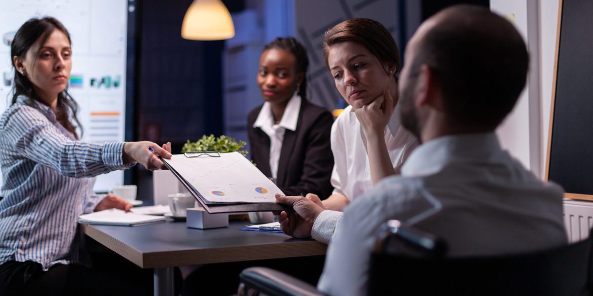 Workaholic disabled leader in wheelchair discussing management statistics solving company graphs. Diverse multi-ethnic businesspeople overworking in meeting room in evening