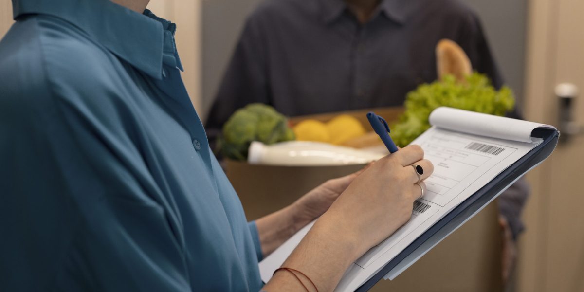 Pessoa verificando checklist em prancheta com caixa de alimentos ao fundo, simbolizando controle e monitoramento de conformidade.