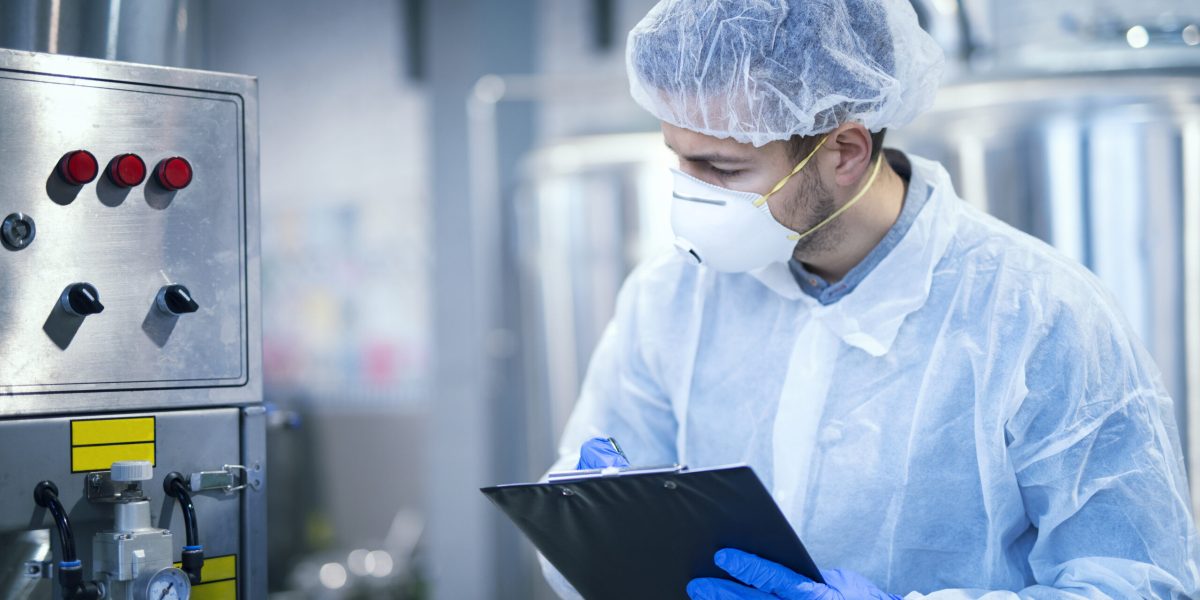 Technologist expert in protective uniform with hairnet and mask taking parameters from industrial machine in food production plant.