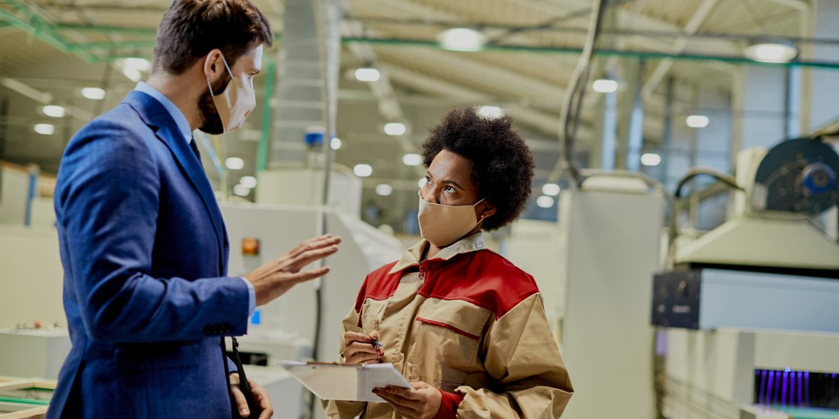Black female worker taking notes while communicating with a businessman who is visiting woodworking production facility during coronavirus pandemic.
